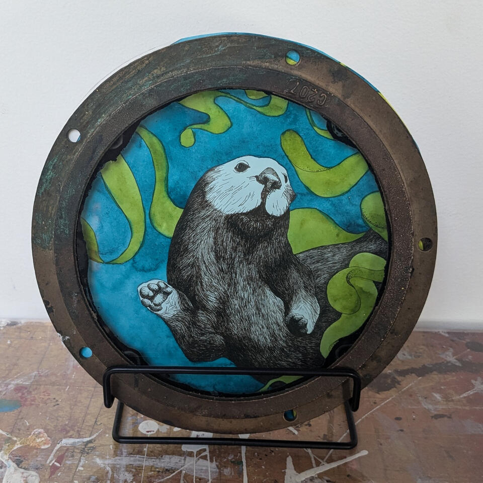 A vintage porthole containing an ink and watercolor image of a sea otter