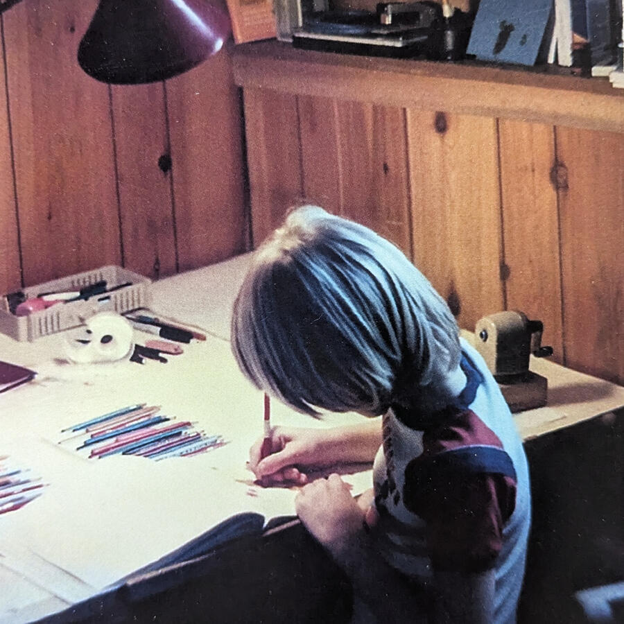 Small blonde child sitting at a table doing art with colored pencils.